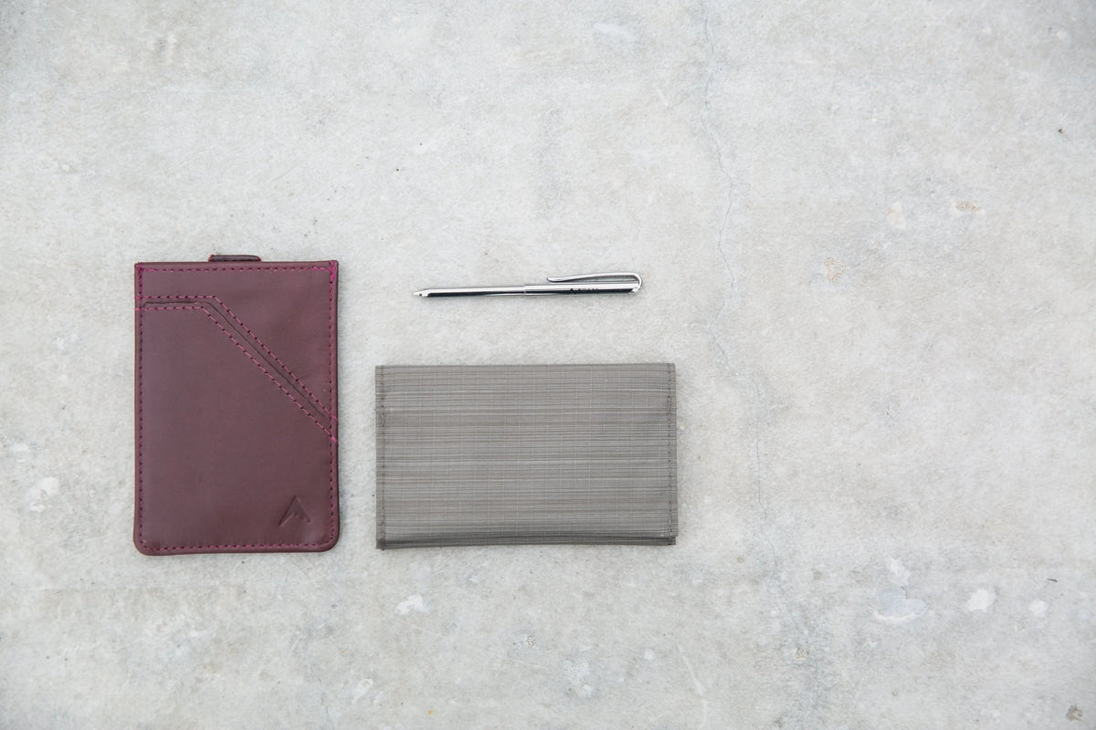 On the left is a maroon leather wallet. On the right is a light grey vegan nylon wallet. Above the vegan wallet is a micropen.