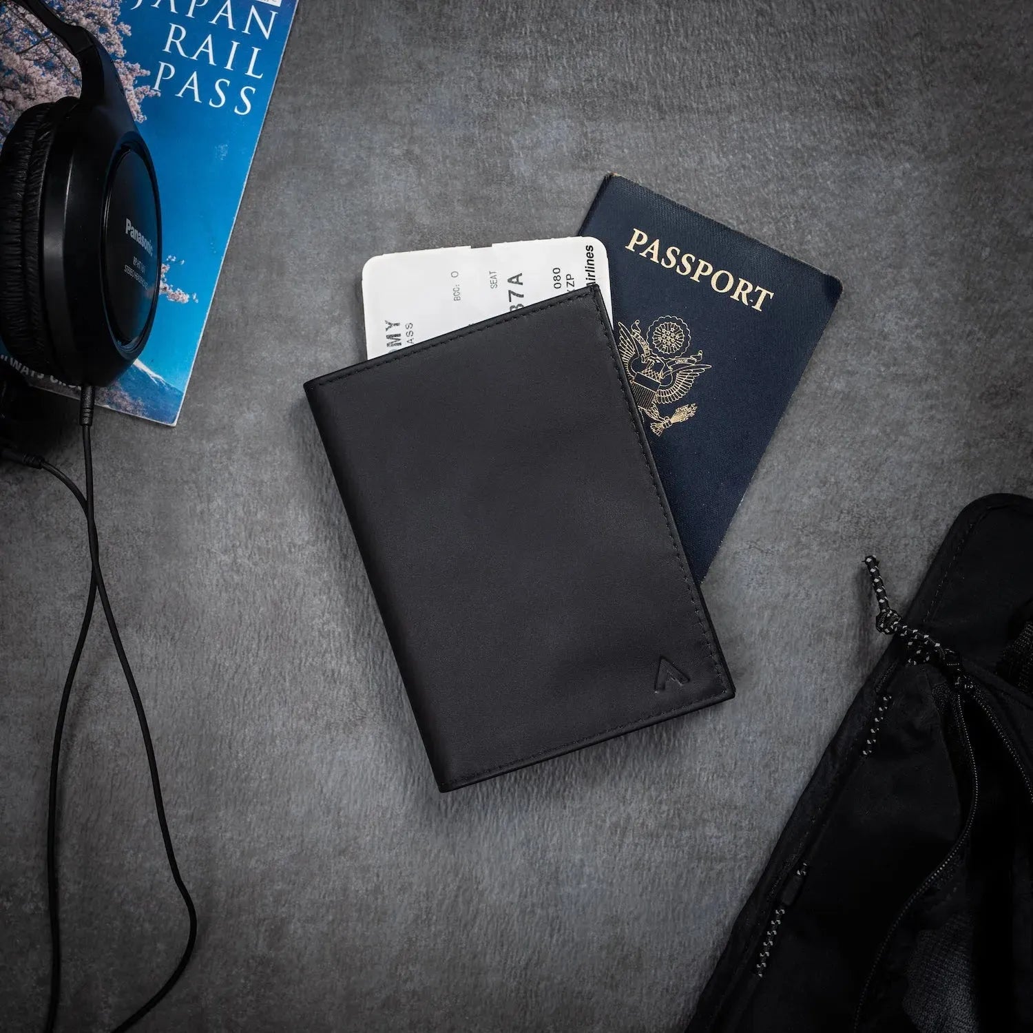 Black leather passport wallet by Allett against a grey table. Underneath the wallet is a passport, and there is also headphones on the table  - black
